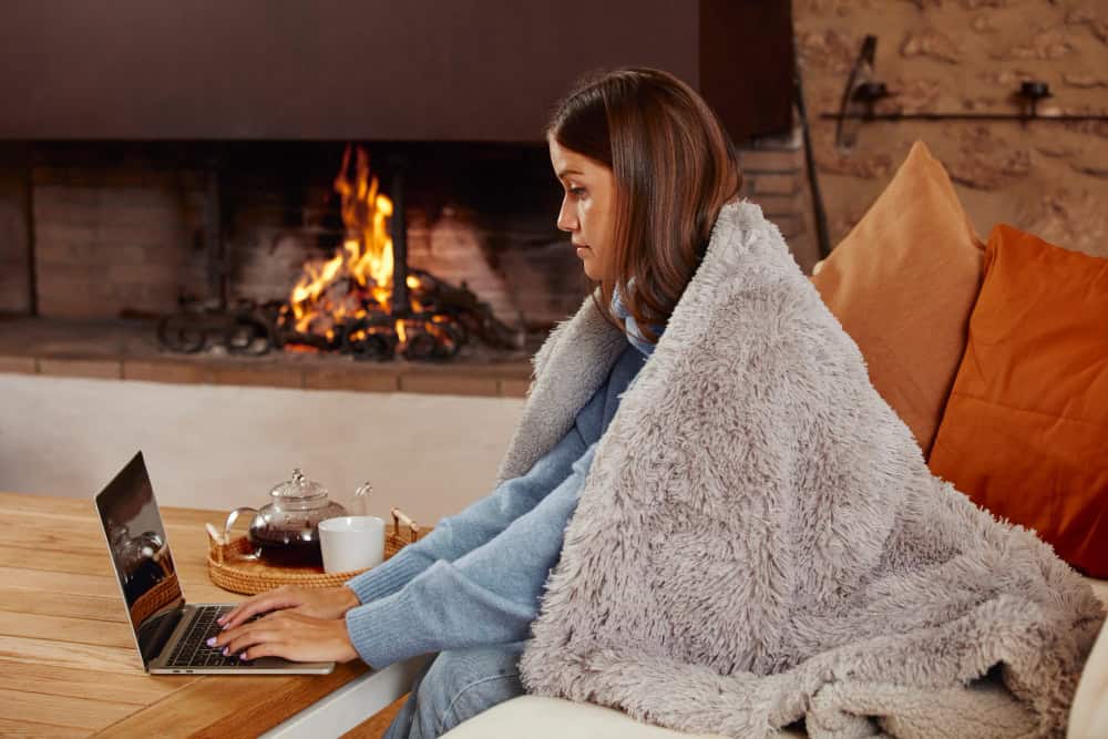 woman wrapped in a grey blanket working at a laptop in front of a roaring fire, with a tea tray beside her
