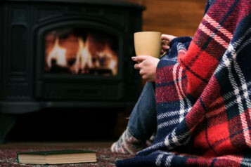 person sitting wrapped in a tartan blanket in front of a wood-burning stove