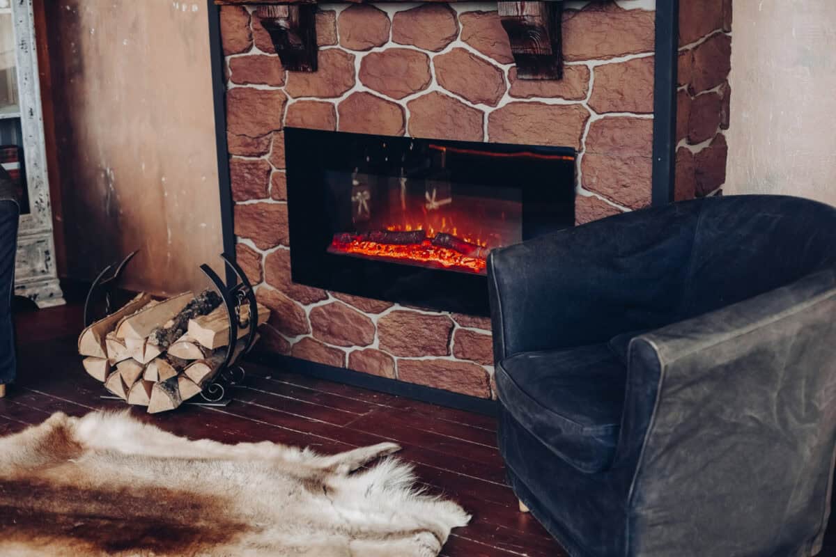 Armchair, fireplace and logs stacked in a round holder, with a fur rug in front of the fire