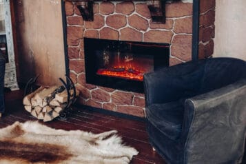 Armchair, fireplace and logs stacked in a round holder, with a fur rug in front of the fire