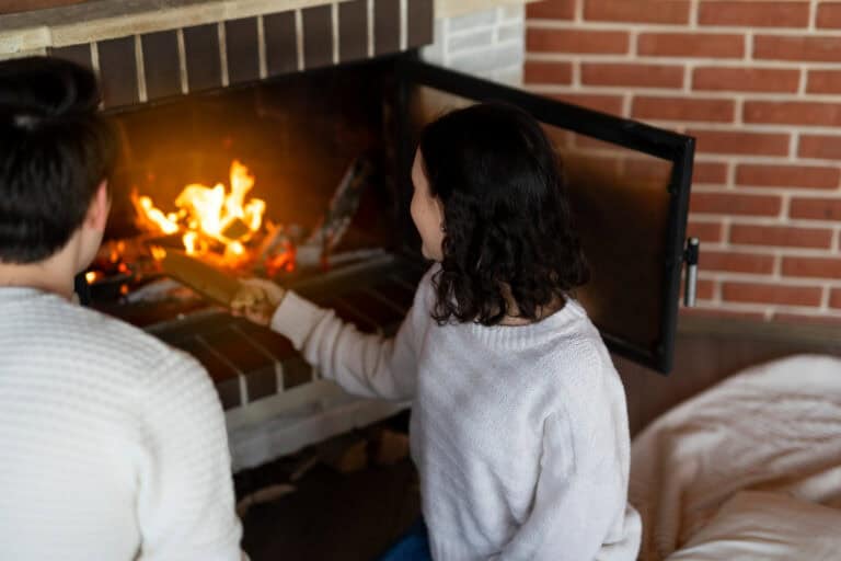 A man and a woman in front of a fireplace, the woman is putting a log on the fire
